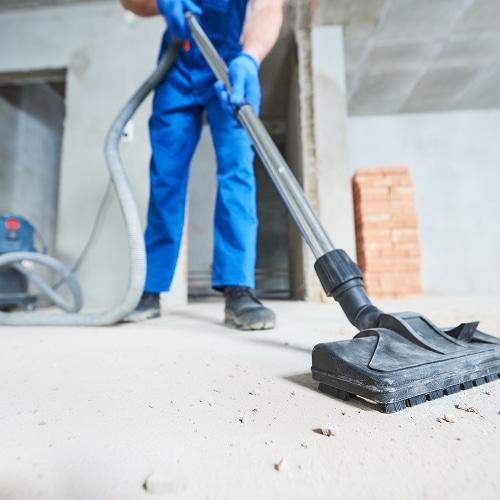 Ouvrier en combinaison bleue et gants, aspirant la poussière sur un sol en béton de chantier avec un aspirateur industriel. Débris visibles.