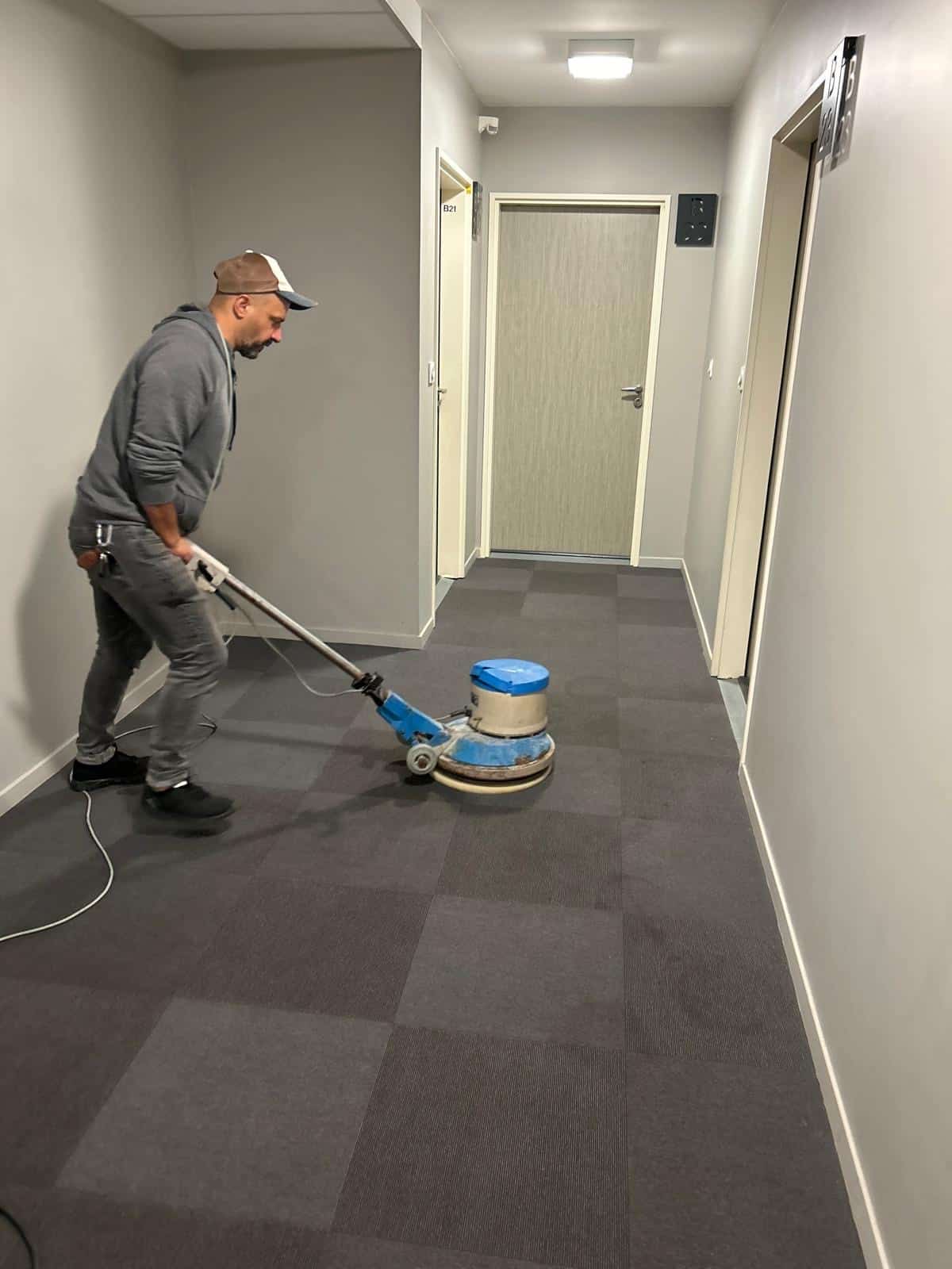 Homme nettoyant un couloir avec moquette foncée à l'aide d'une machine professionnelle. Porte B21 visible. Propreté des sols.